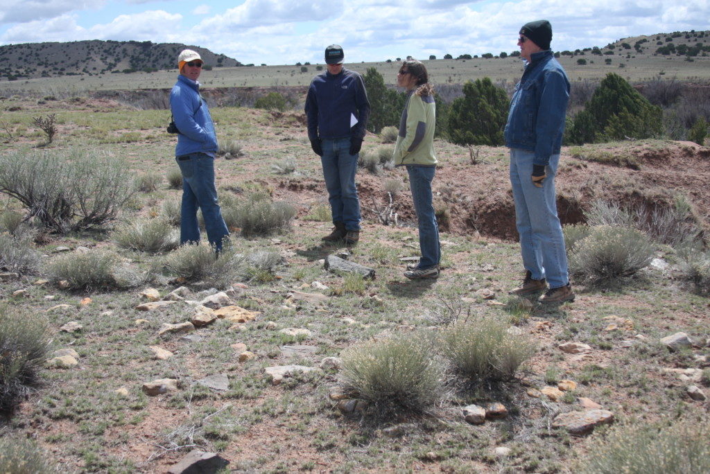 Photo of three men and one woman standing in a sparsely vegetated field with a large dropoff into an arroyo just bahind them. Cloudy sky in background.