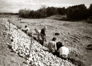 Black and white photo of people arranging rocks and stabilization material along the edge of an arroyo.