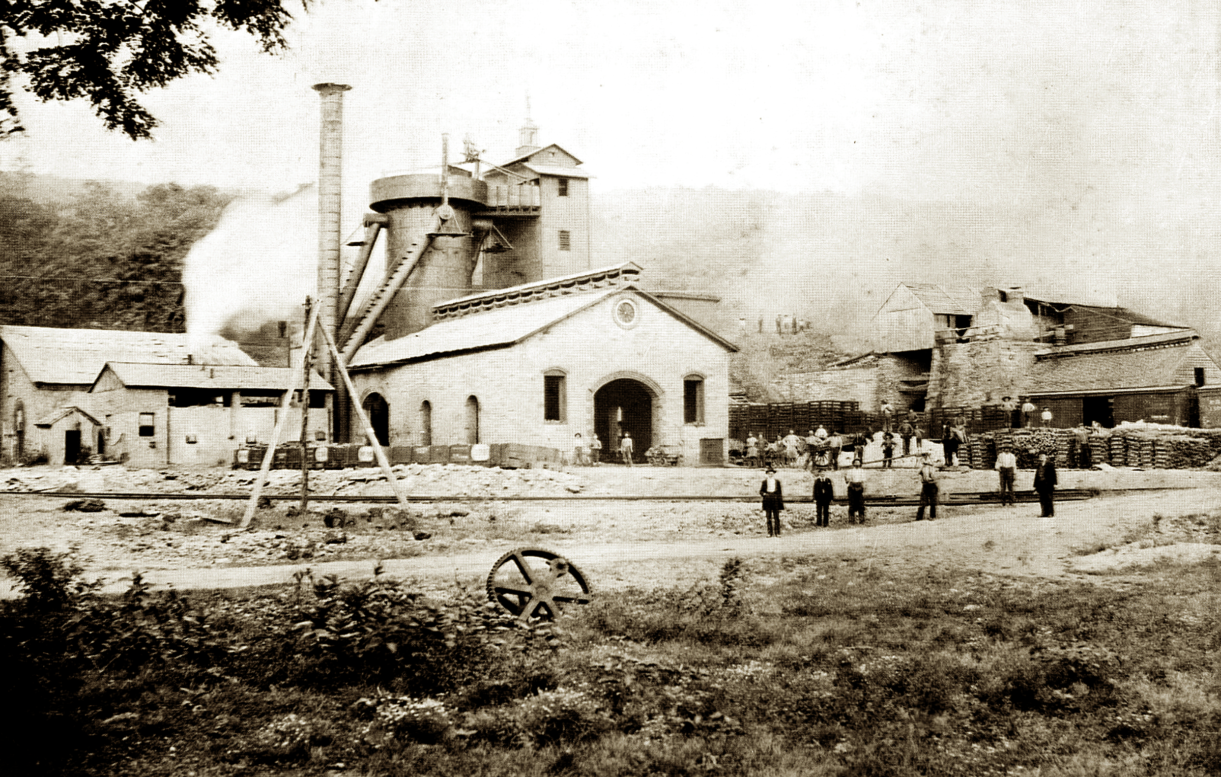 Black and white photo showin a white building with large central doorway. Associated buildings lie to the left and right, and a tall smokestack and tower are attached toward the rear of the building.