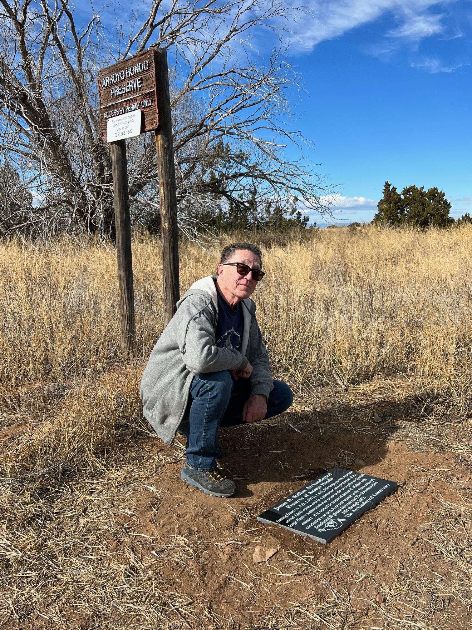 Photo of man kneeling in front of plaque installed in the ground. Blue sky in background with leafless trees and a wooden sign.