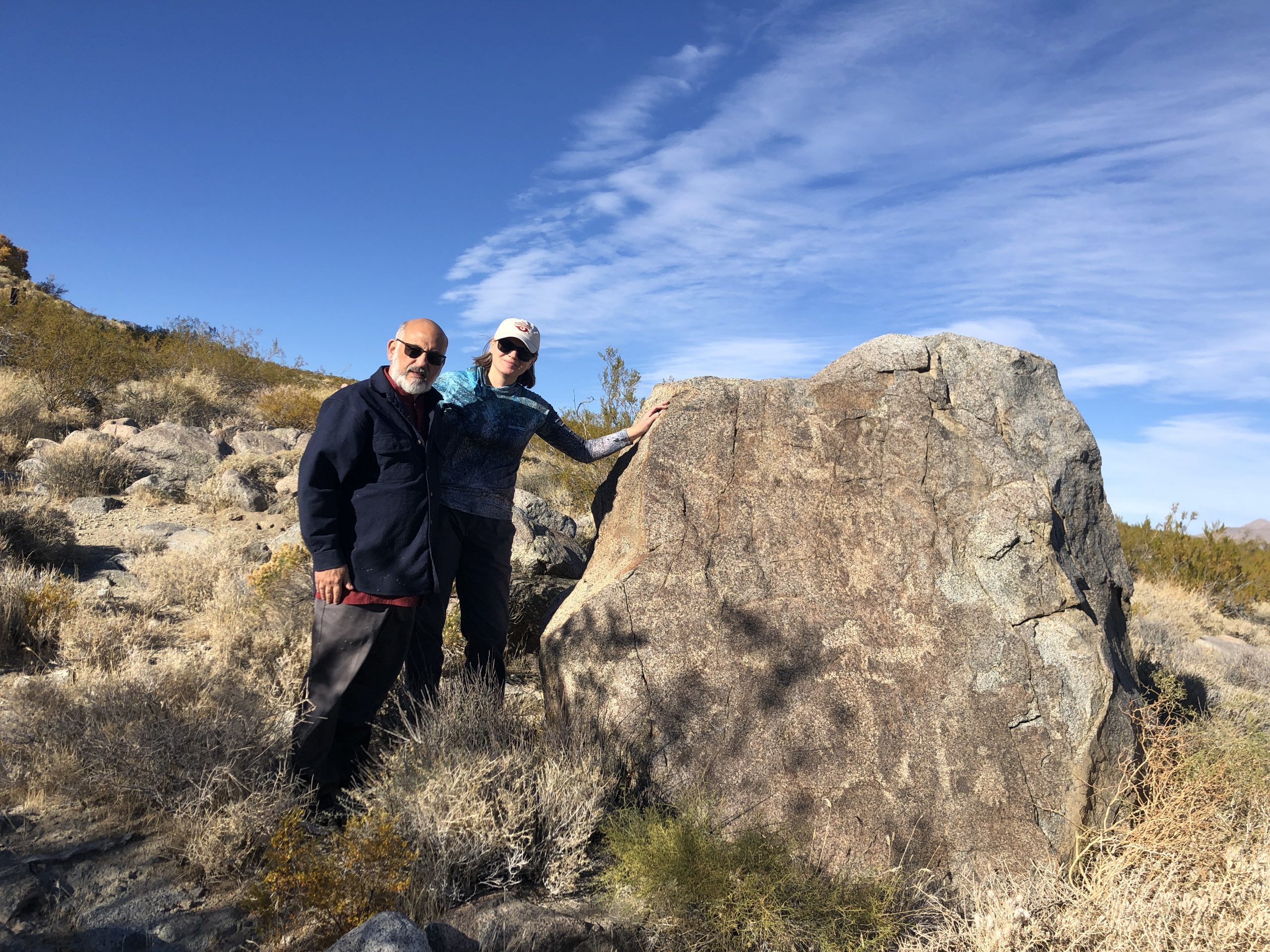 A man and a woman stand near a petroglyph-covered boulder in a desert setting.