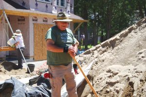 photo of man wearing hat and holding a tool with a long wooden handle in what appears to be a construction site.