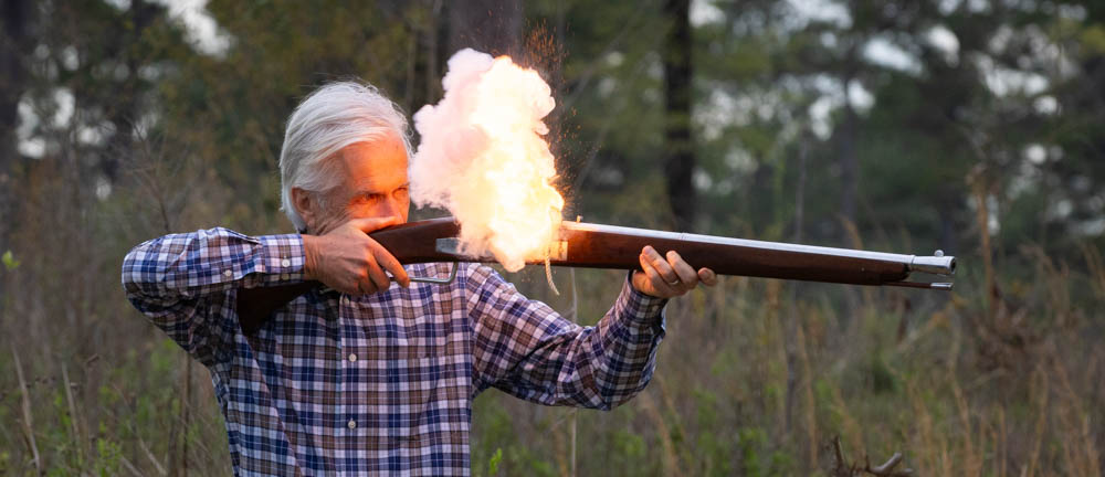 Photo of an older man shooting a replica musket with explosive fire coming up from the center of the gun.