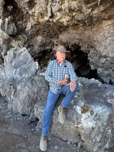 Photo of a man wearing a plaid shirt, denim jeans, and a hat. He is leaning up agains a rock formation with a cave in the background.