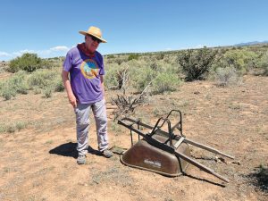 Man in purple shirt and straw hat stands next to an abandoned upsidedown wheelbarrow.