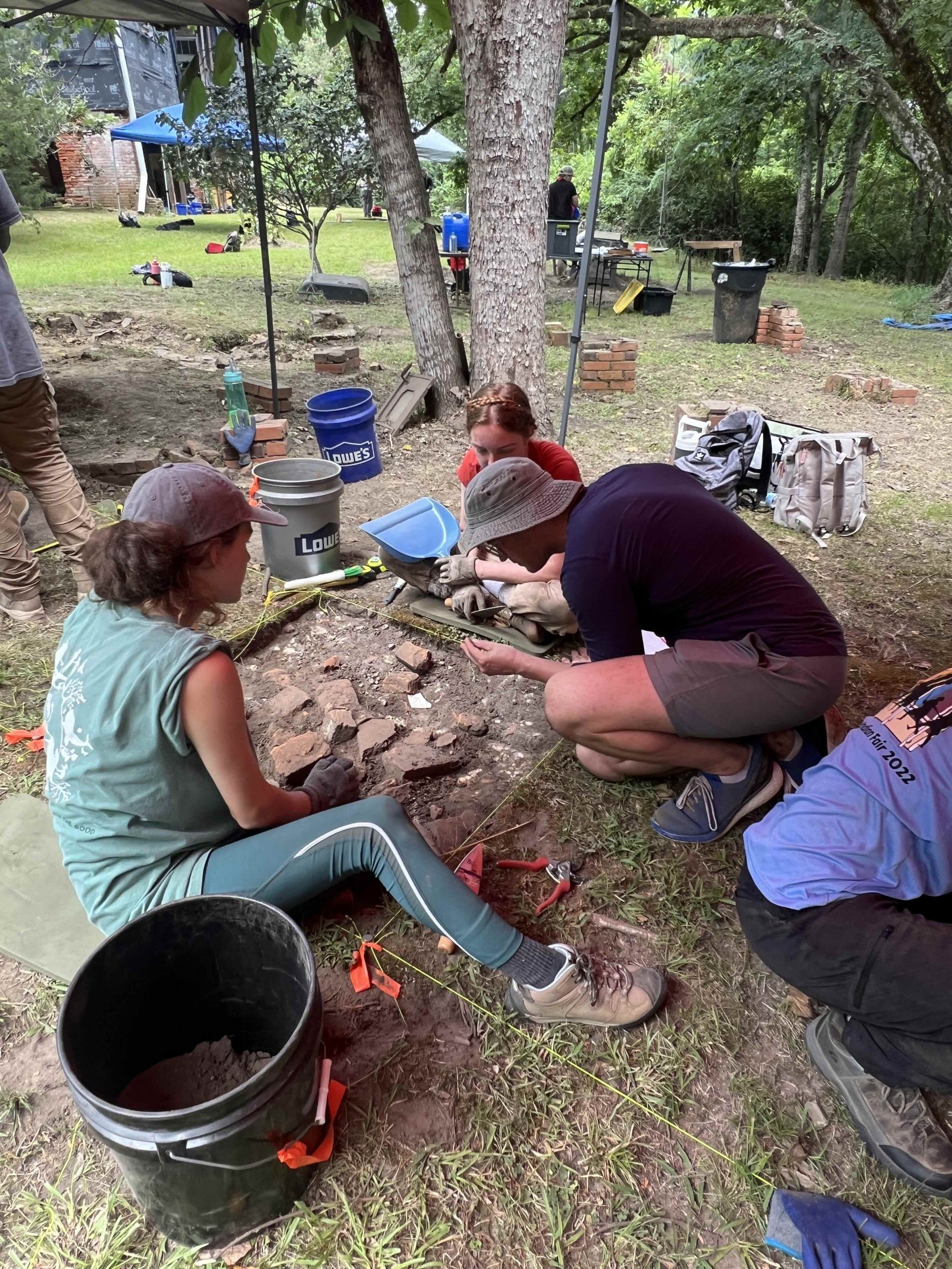 A man kneeling in front of a excavated area is holding an artifact while several others look on.